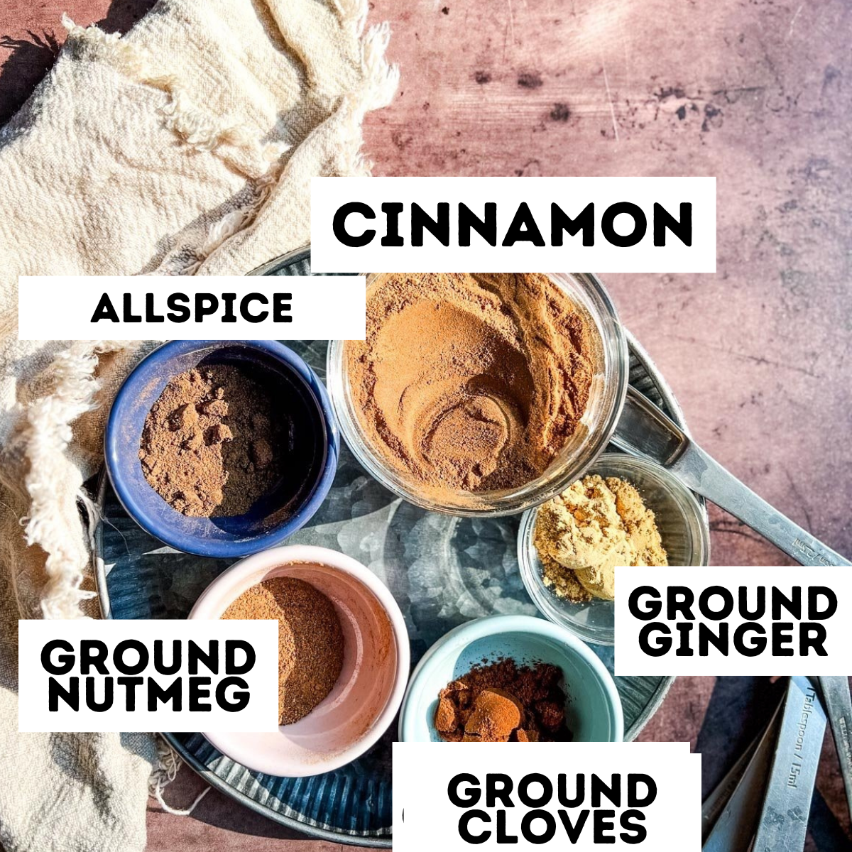 A plate with bowls of cinnamon, allspice, nutmeg, cloves, and ginger on a rustic pink surface.