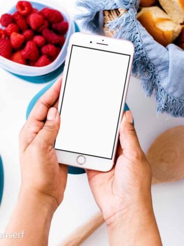 Person holding a blank phone over a table with raspberries, rolls, Brussels sprouts, and a wooden spoon.