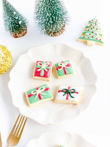 Iced Christmas cookies shaped like gift boxes on a white plate, with small trees as decoration.