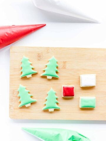 Christmas tree and gift-shaped cookies on a wooden board with red, green, and white icing piping bags.