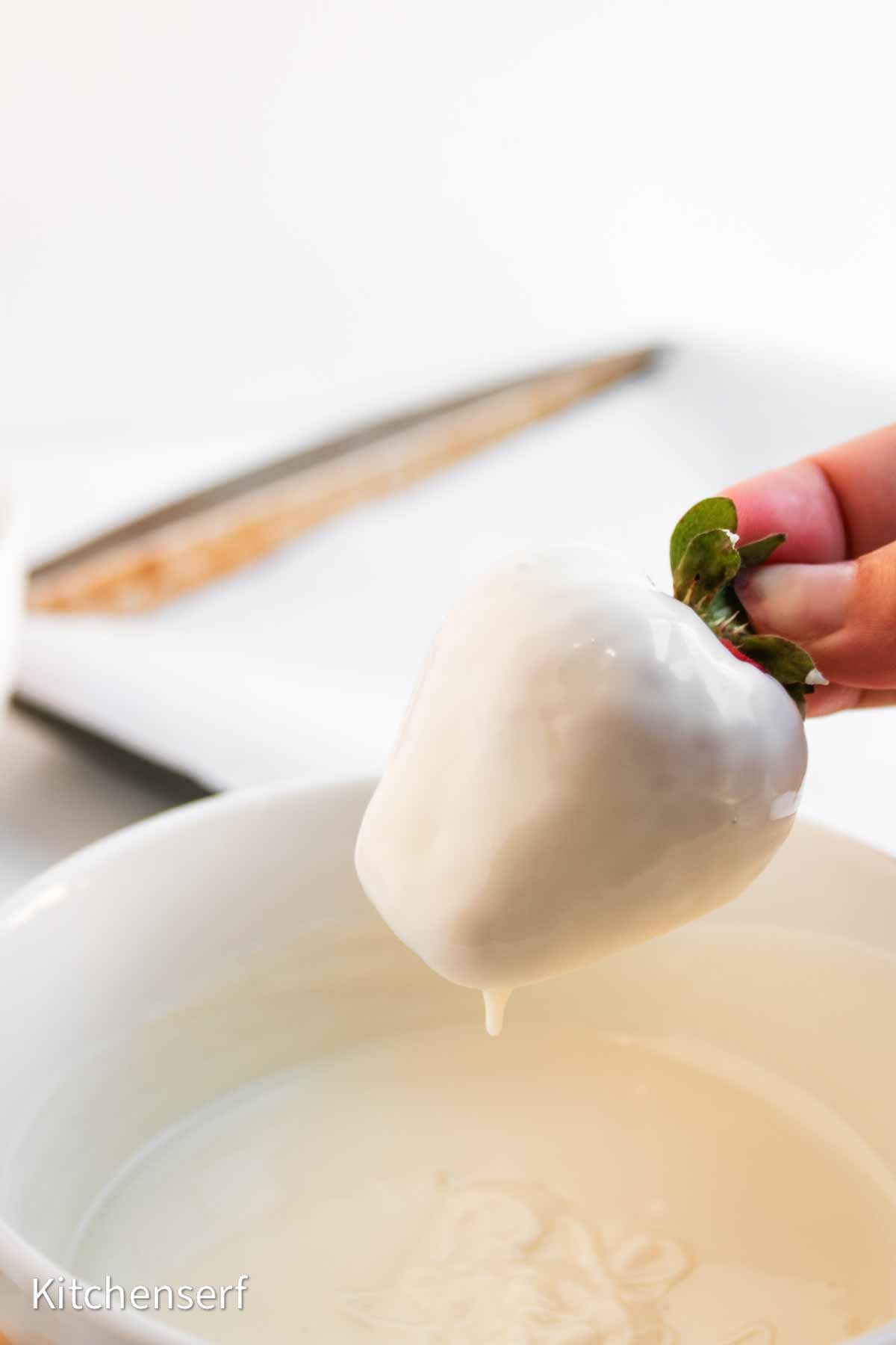 A hand holds a strawberry dipped in white chocolate above a bowl of melted white chocolate.