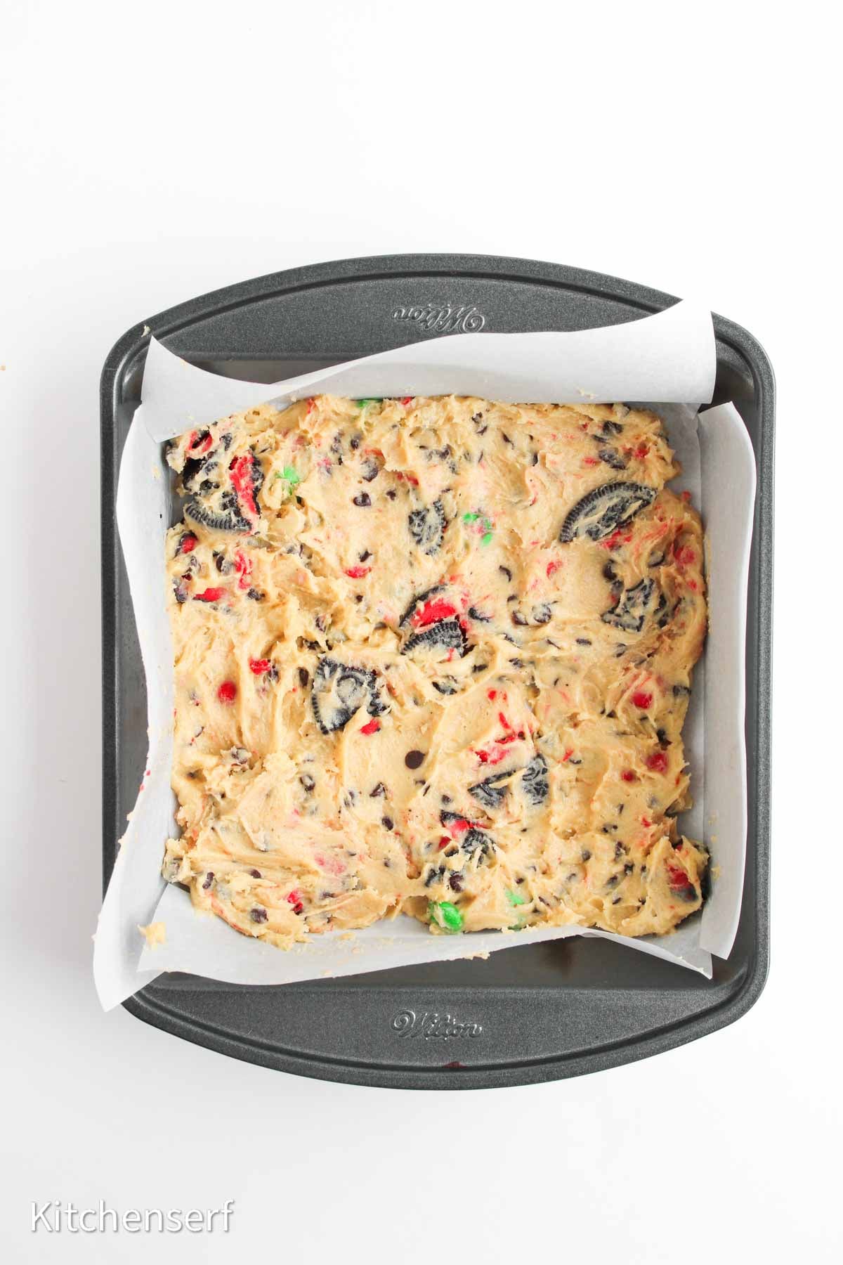 Square baking pan filled with cookie dough mixed with crushed cookies and colorful candy pieces, on parchment paper.