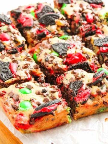 Nine Christmas cookie bars topped with Oreo pieces, red and green candies, and chocolate chips on parchment paper.
