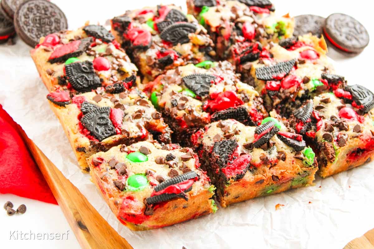 Nine Christmas cookie bars topped with Oreo pieces, red and green candies, and chocolate chips on parchment paper.
