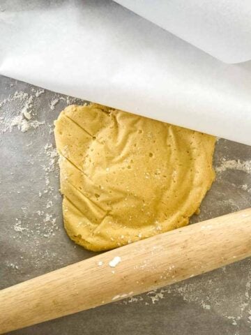 Rolled sugar cookie dough between sheets of parchment paper with a rolling pin nearby, perfect for cut-out cookie troubleshooting.