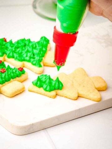 Hand piping green icing onto Christmas tree-shaped cookies on a white surface.