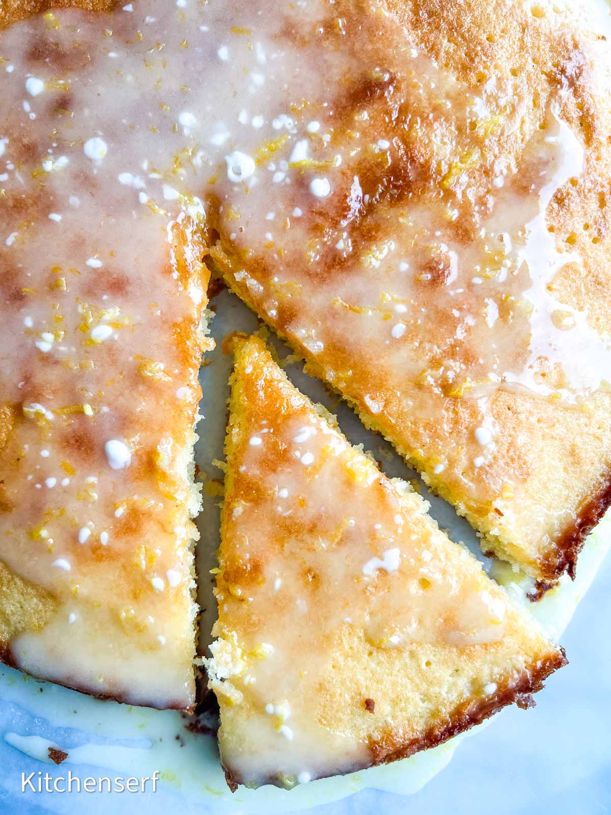 A glazed lemon cake with one slice partially removed, showing a moist, golden interior.