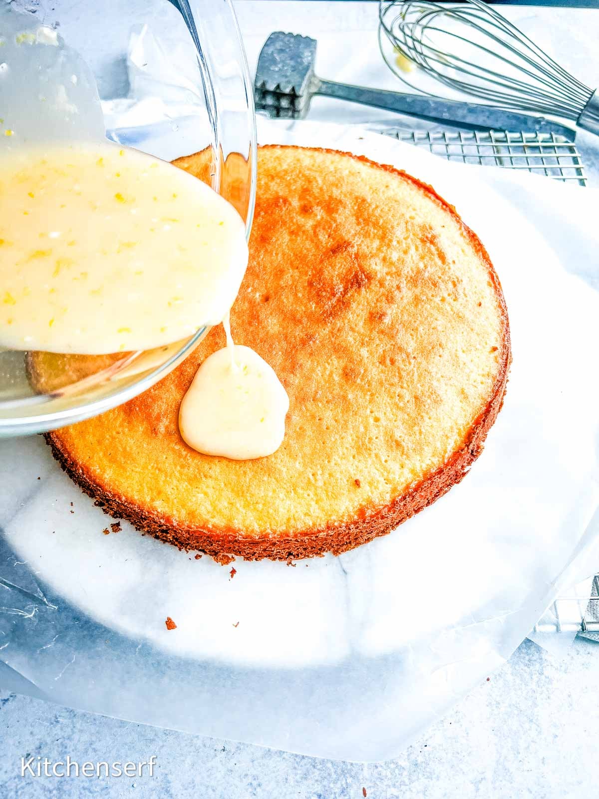 Lemon glaze being poured onto a round yellow cake on parchment paper with baking tools nearby.