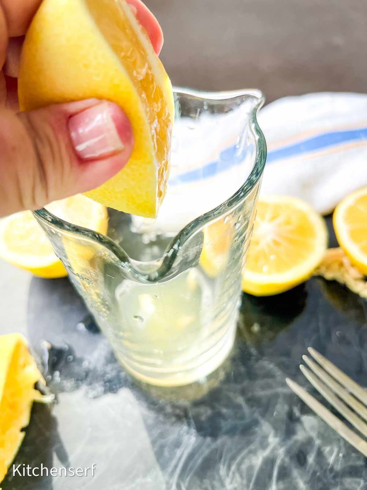 Hand squeezing a lemon wedge over a glass pitcher with halved lemons and a fork on a table.