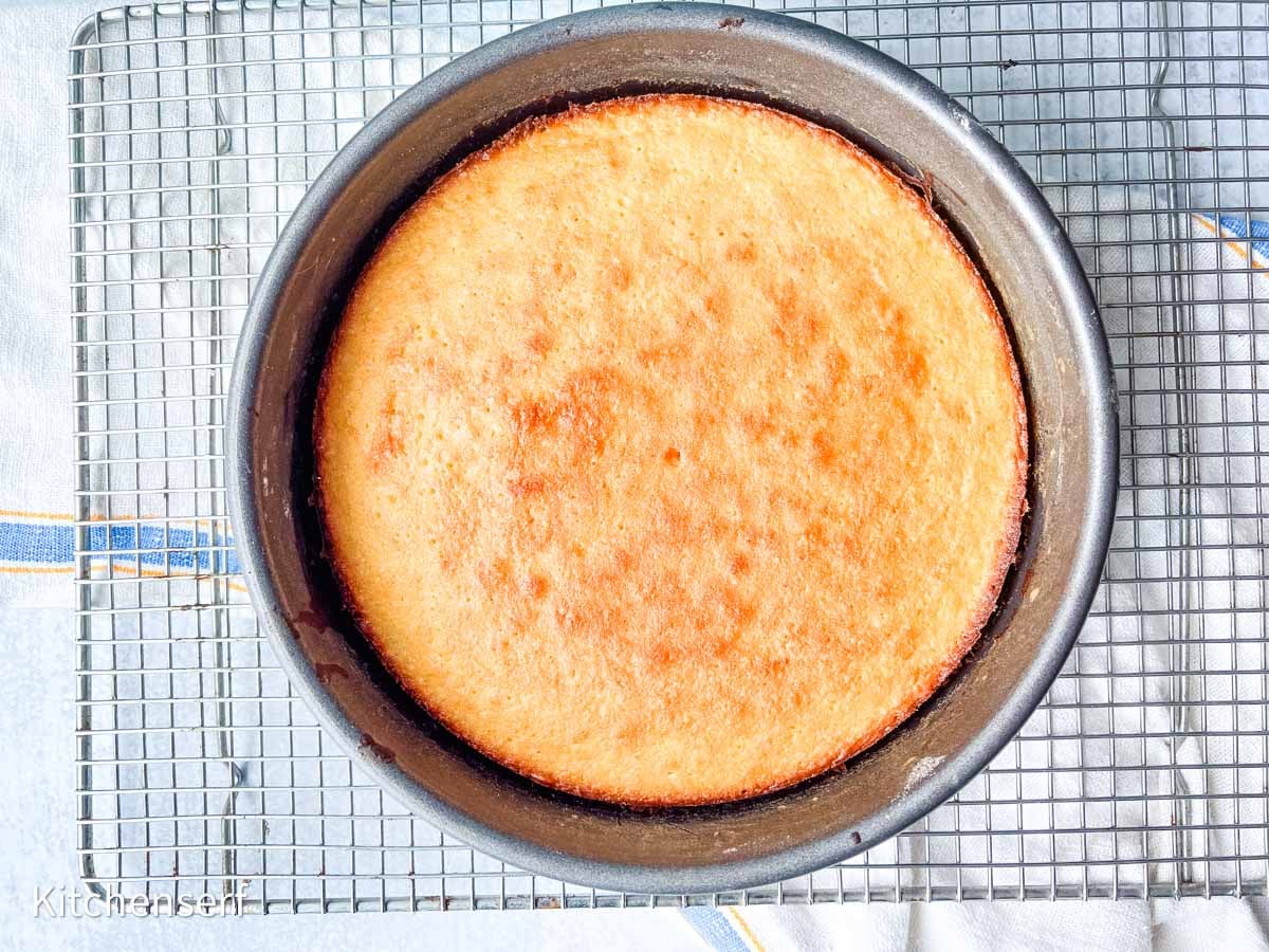 A round, golden-brown cake in a baking pan rests on a cooling rack over a white towel.