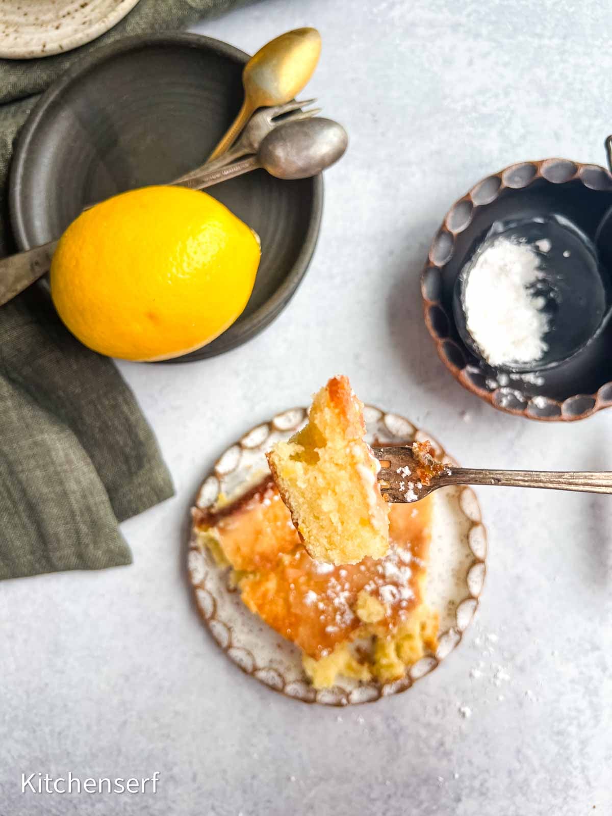 A fork holds a bite of meyer lemon cake above a plate, with a whole lemon and bowls nearby.