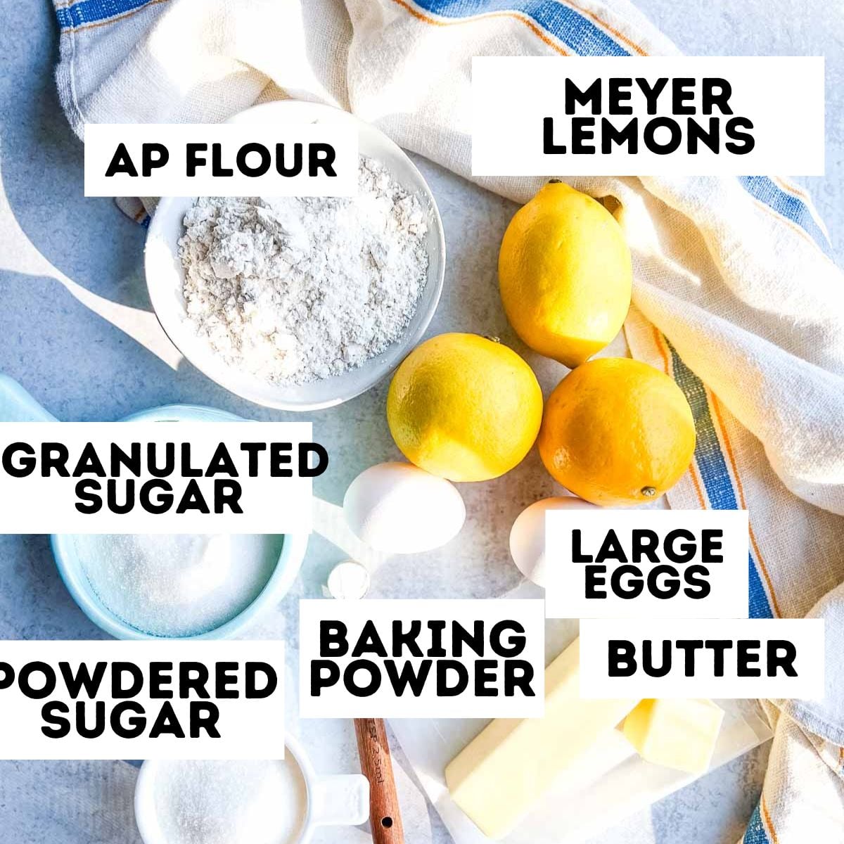 Flour, sugar, eggs, butter, Meyer lemons, and baking powder labeled on a kitchen counter with a towel.