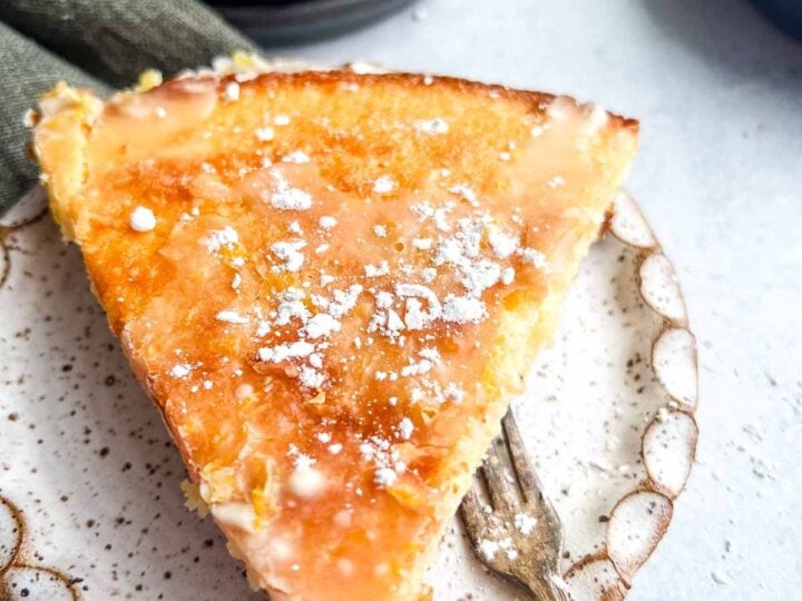 A slice of glazed Meyer lemon cake on a speckled plate with a fork, sprinkled with powdered sugar.