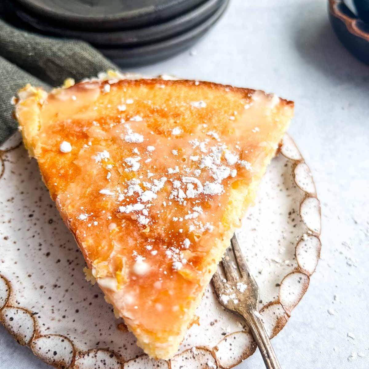 A slice of glazed Meyer lemon cake on a speckled plate with a fork, sprinkled with powdered sugar.