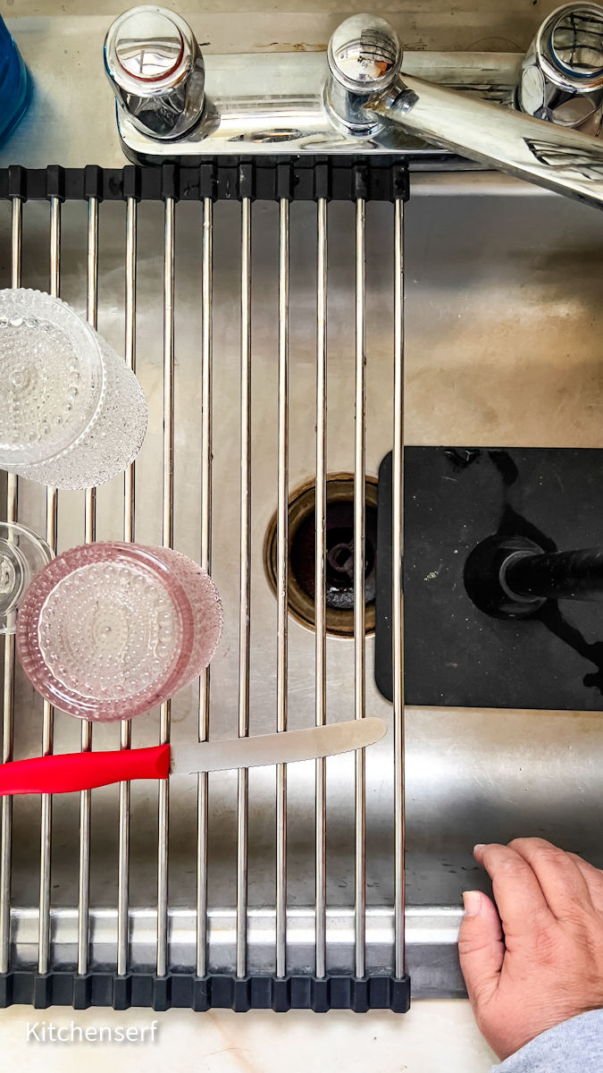 A hand near a sink with glasses, a knife, and a drying rack over the basin.