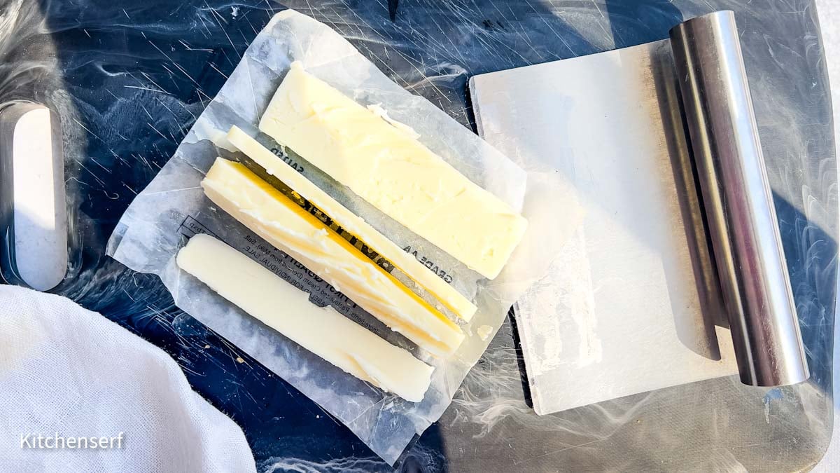 Three sticks of butter on wax paper next to a metal bench scraper on a dark countertop.