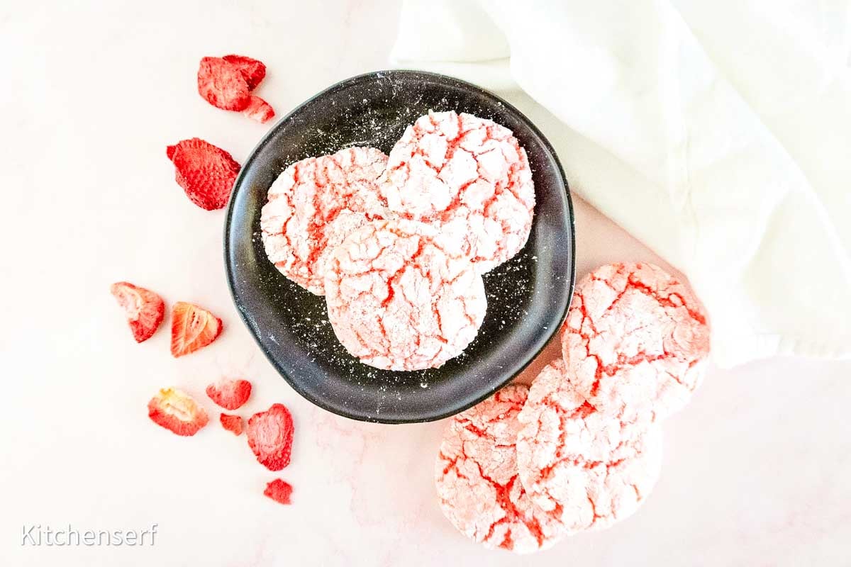 Pink crinkle cookies on a black plate, with dried strawberries and more cookies on a light surface nearby.