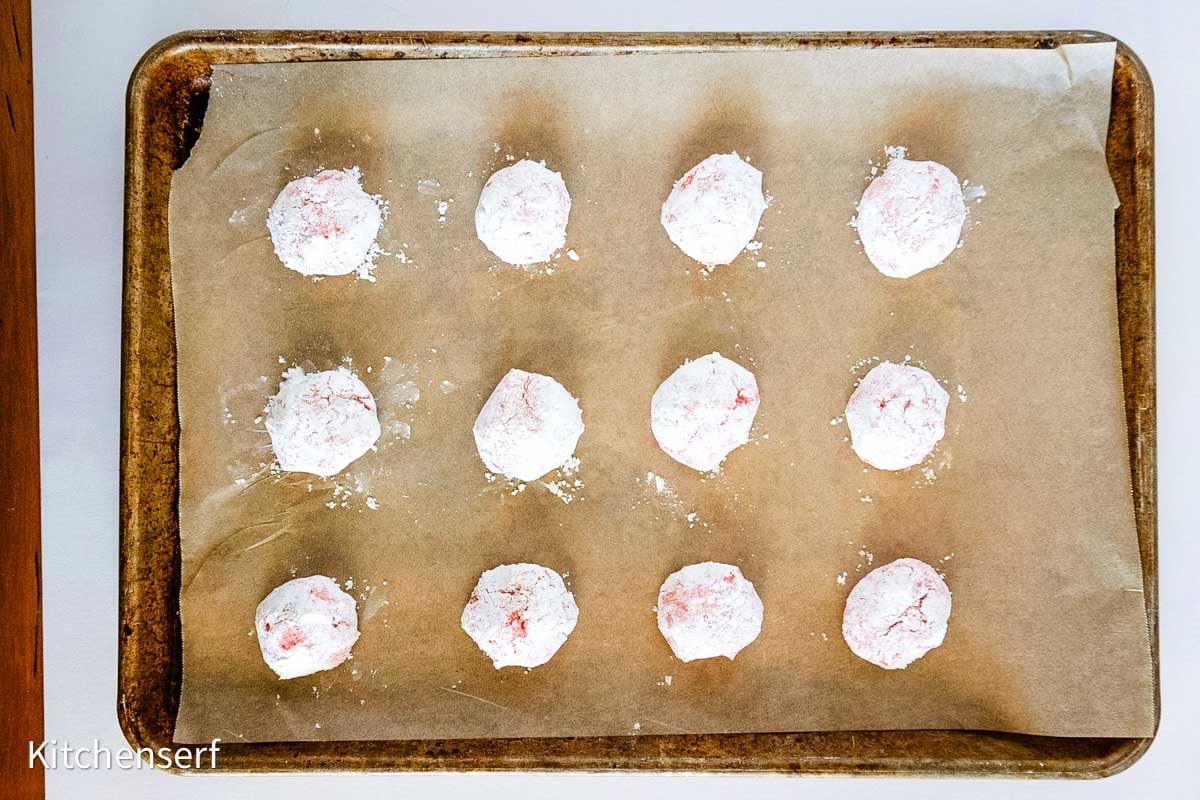 Twelve powdered sugar-coated cookie dough balls on a parchment-lined baking sheet, ready to bake.