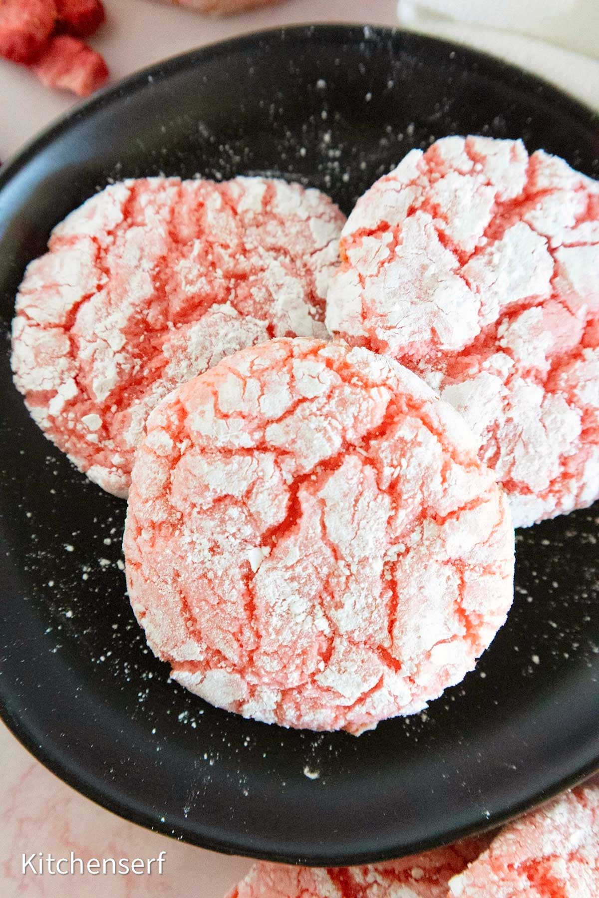 Three pink crinkle cookies coated in powdered sugar on a black plate.
