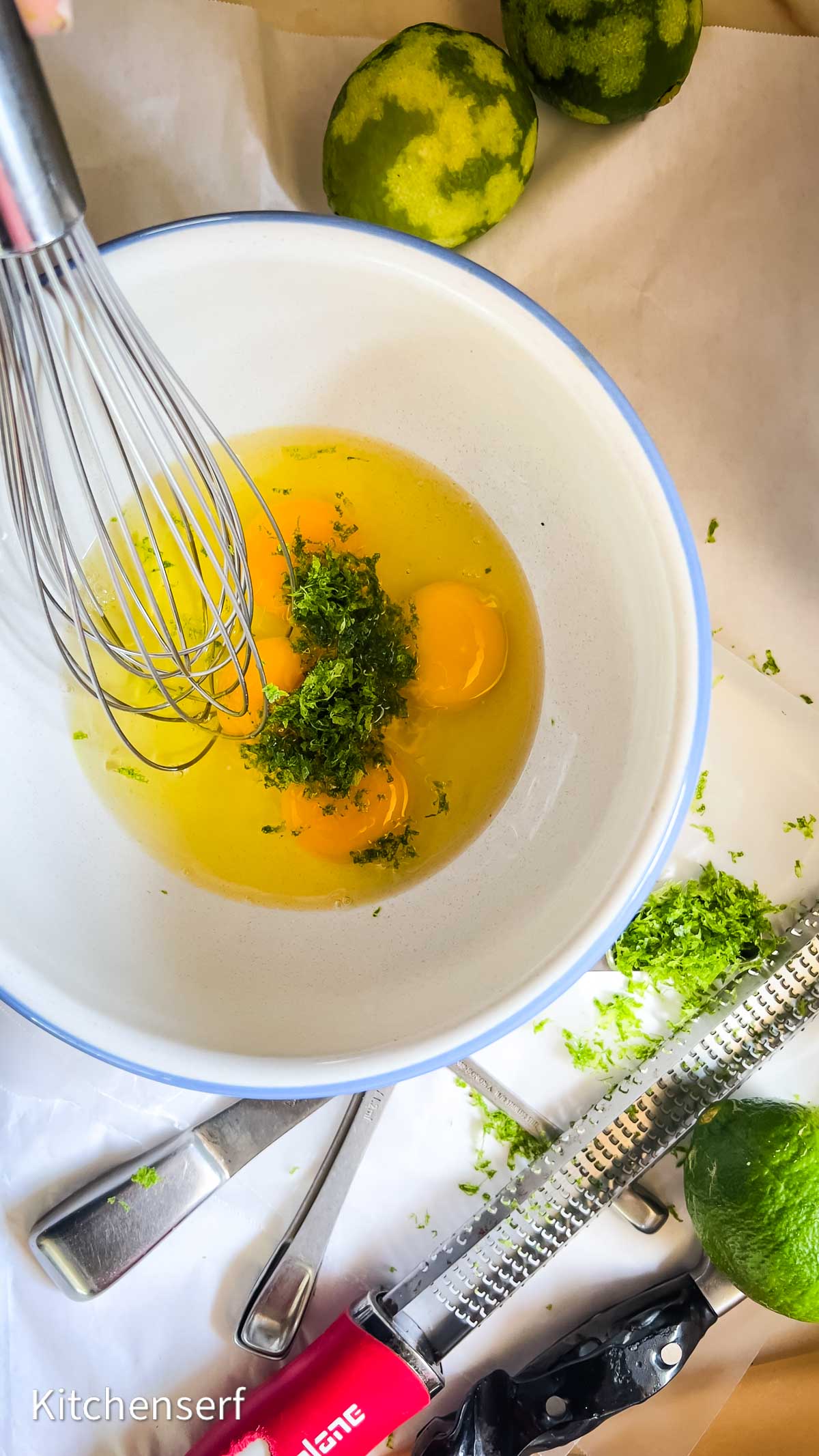 Three eggs and lime zest in a bowl with a whisk, surrounded by limes and a grater on a white surface—ingredients for starting a classic key lime pie.