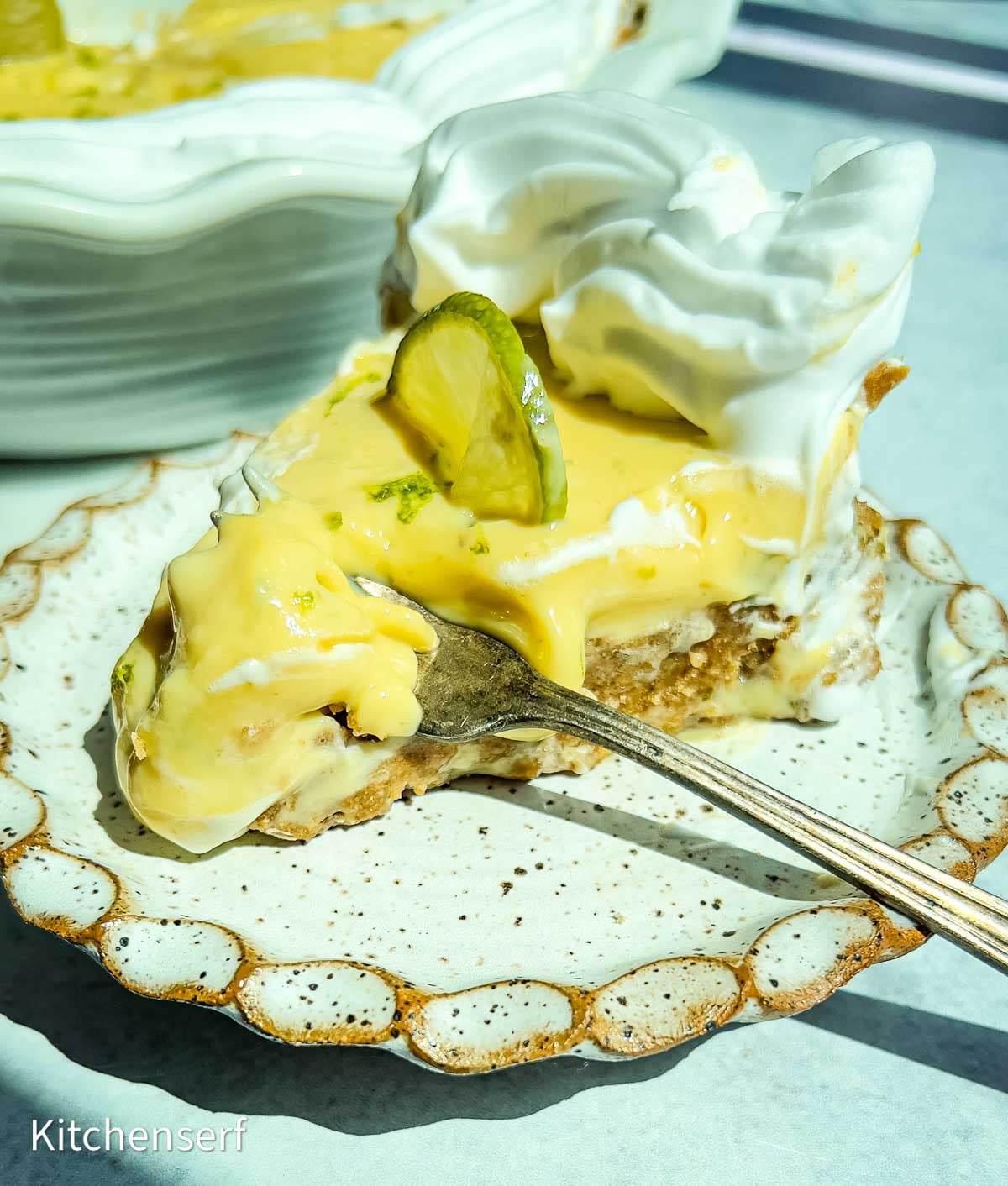 A slice of creamy pie with whipped topping, a lime slice on top, and a fork on a decorative plate.