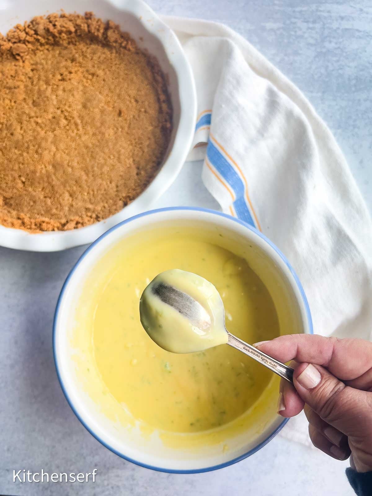 Hand holding a spoon of yellow classic key lime pie filling over a bowl, with a graham cracker crust and towel nearby.