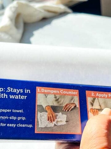 Hand holding a box of countertop prep paper, showing usage instructions and non-slip lining feature near a sunny window.