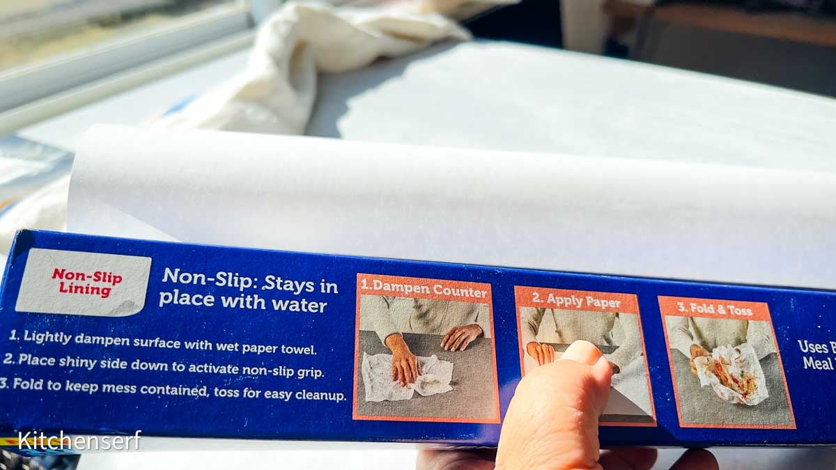 Hand holding a box of countertop prep paper, showing usage instructions and non-slip lining feature near a sunny window.