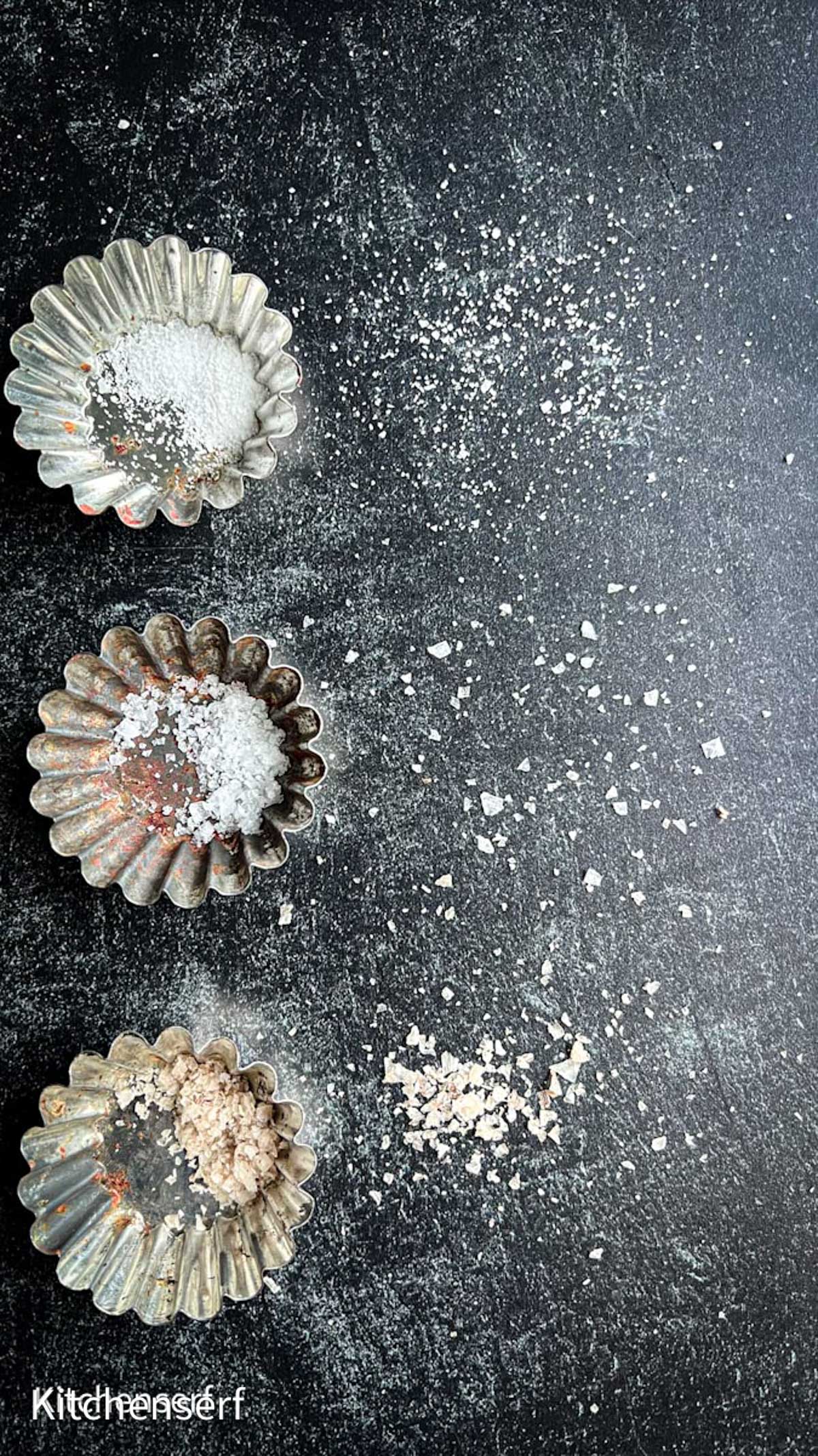 Three small fluted metal dishes hold the best salt for baking cookies, each with a different variety, displayed on a dark, textured surface with salt scattered around.