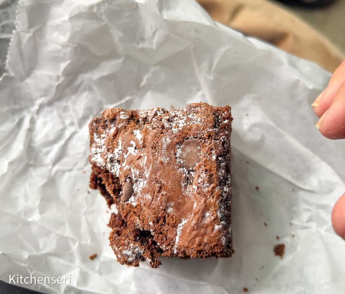 A partially eaten chocolate brownie, made better with boxed brownie mix hacks, is dusted with powdered sugar and sits on a crumpled piece of white parchment paper.