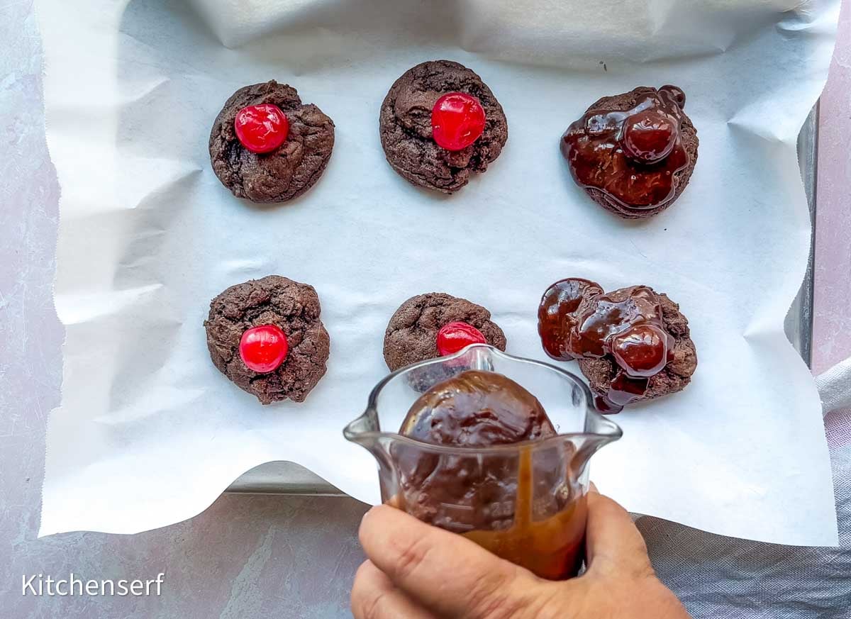 Six chocolate cookies with cherries on parchment; a hand holds glass container to pour chocolate ganache over some of the cookies.