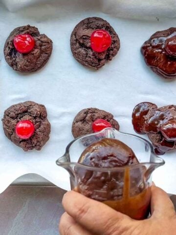 Six chocolate cherry cookies rest on parchment; three are topped with cherries, while a hand drizzles rich chocolate sauce over the others.