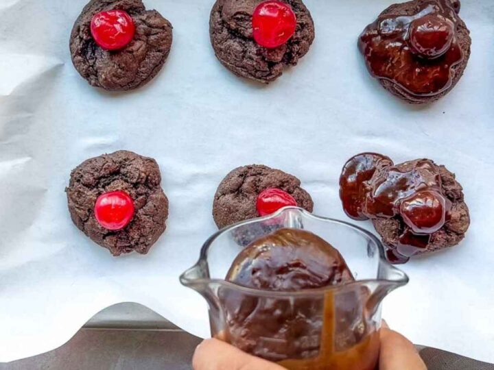Six chocolate cherry cookies rest on parchment; three are topped with cherries, while a hand drizzles rich chocolate sauce over the others.