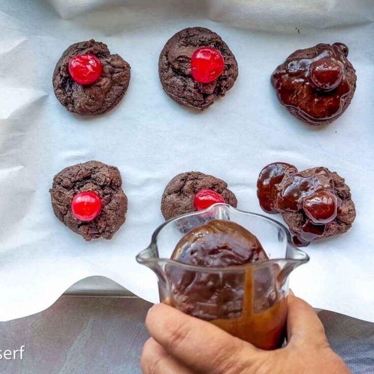Six chocolate cherry cookies rest on parchment; three are topped with cherries, while a hand drizzles rich chocolate sauce over the others.