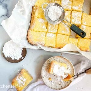 A tray of lemon bars dusted with powdered sugar, one bar served on a plate with extra sugar on top.