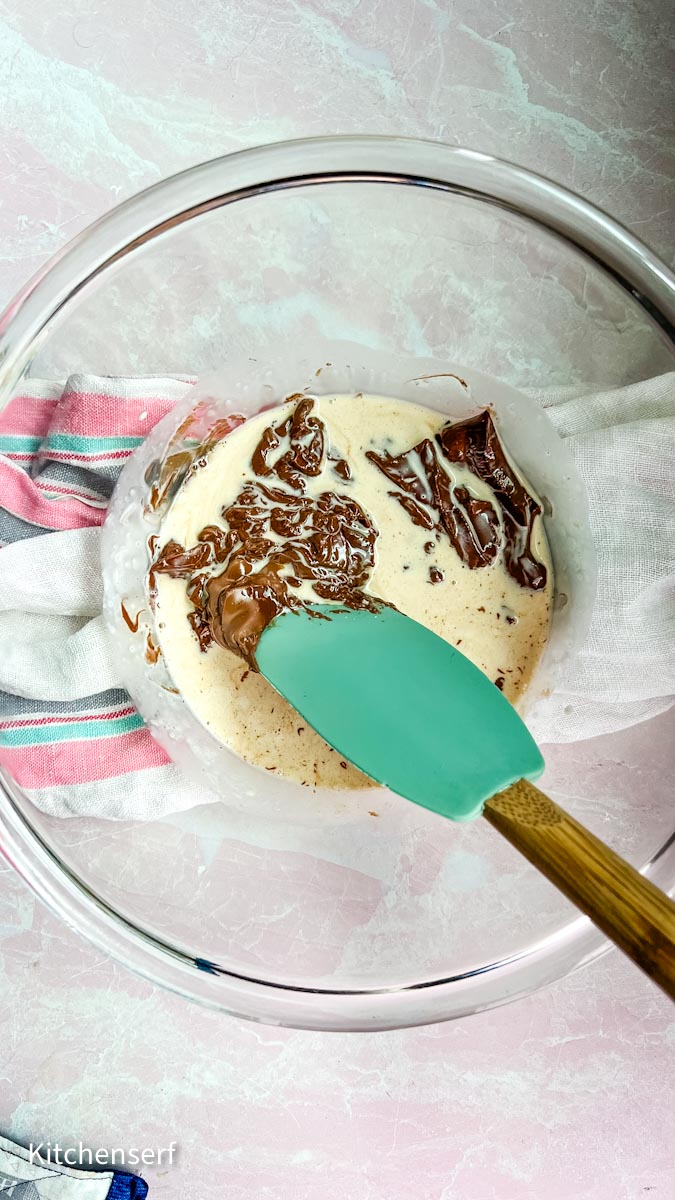 A blue spatula stirring cream and chocolate in a glass bowl, held steady with a striped towel.