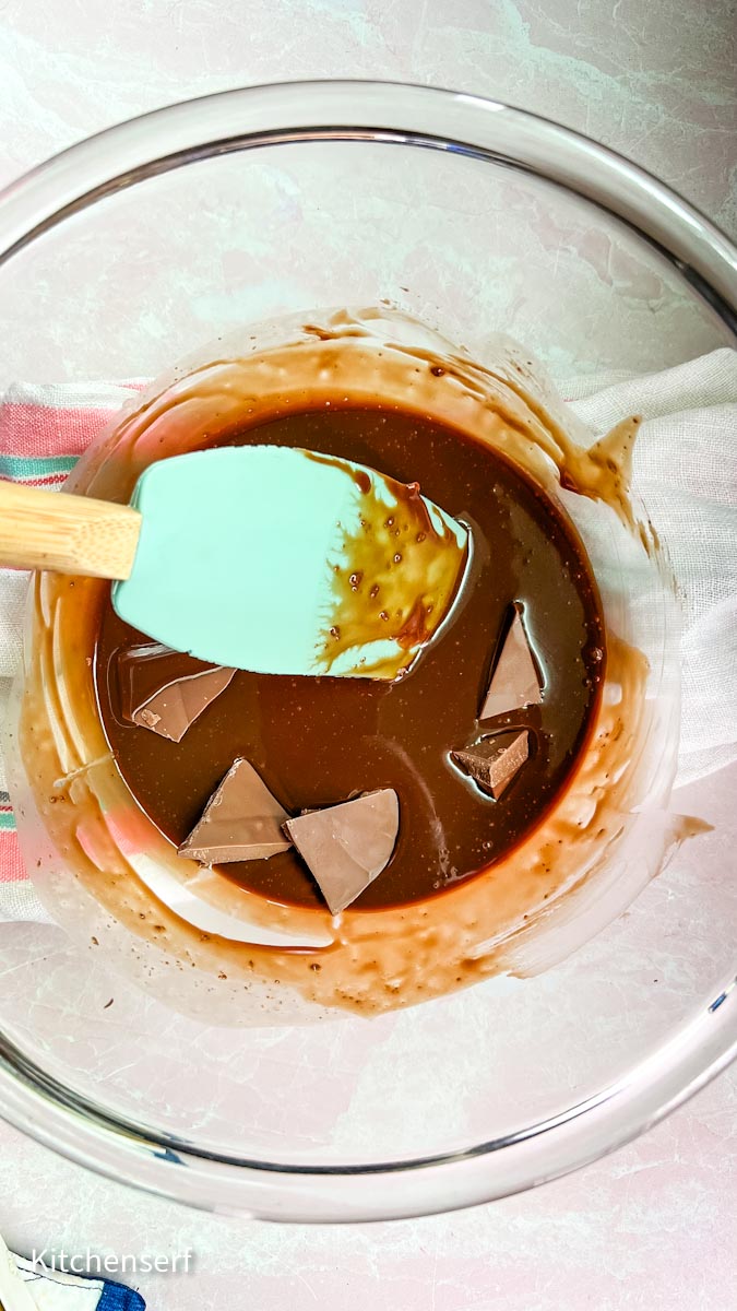 A spatula stirs melting chocolate pieces in a glass bowl on a pink surface.