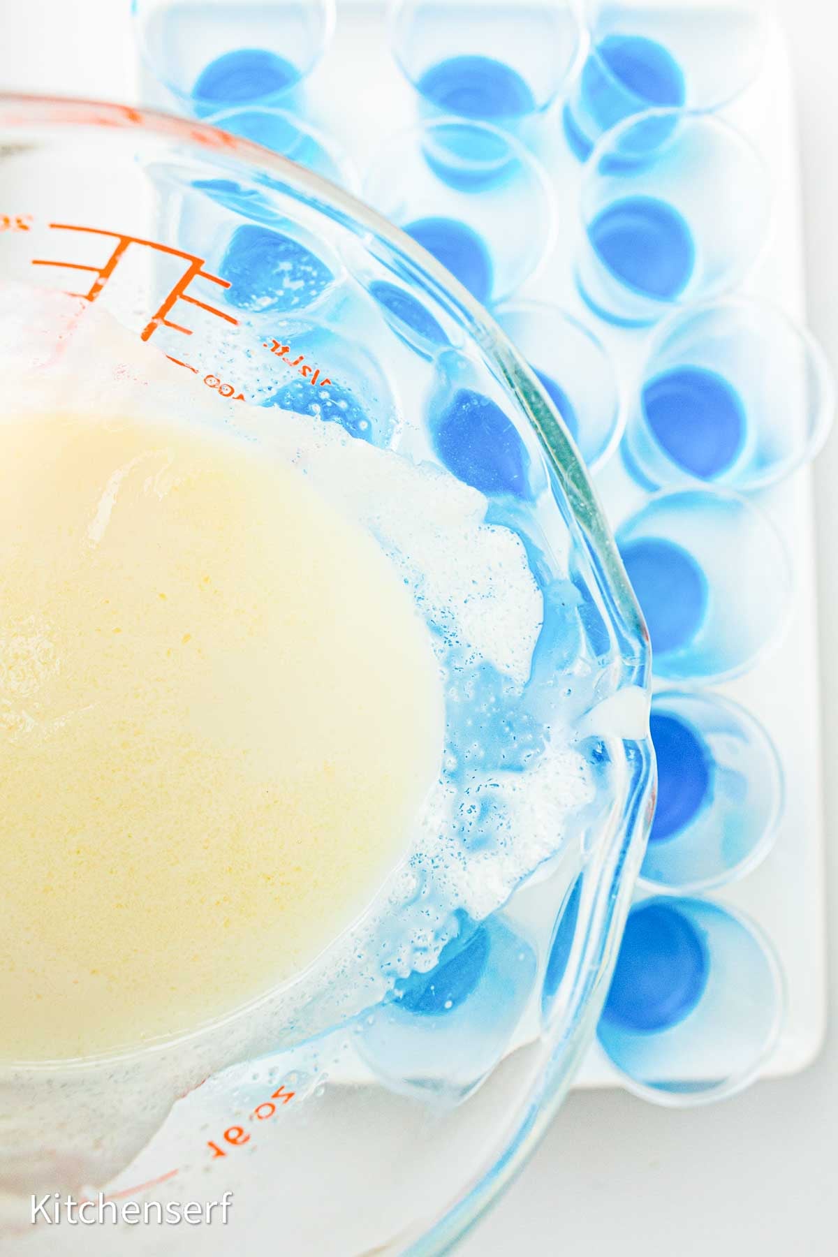 A glass measuring cup with milk is being poured into small blue cups arranged on a white tray.