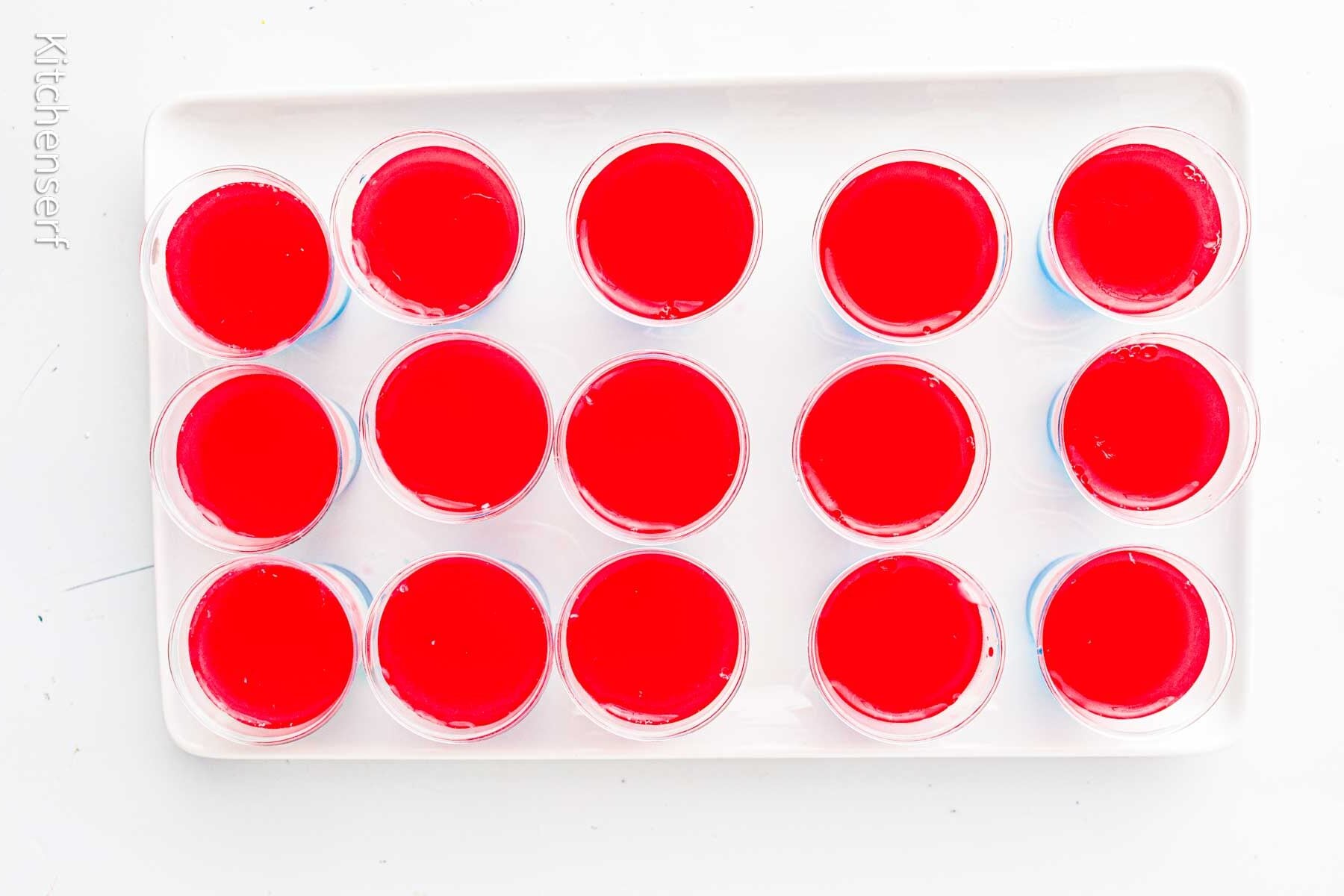 15 red jello shots in clear plastic cups arranged in rows on a white rectangular tray.