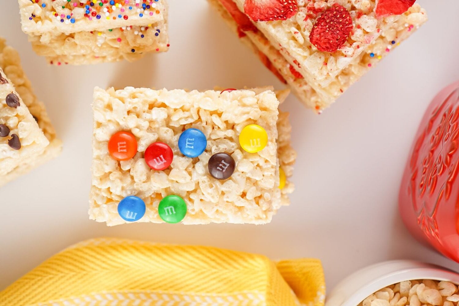 A sheet pan Rice Krispies treat topped with colorful M&Ms sits on a white surface next to other decorated treats.