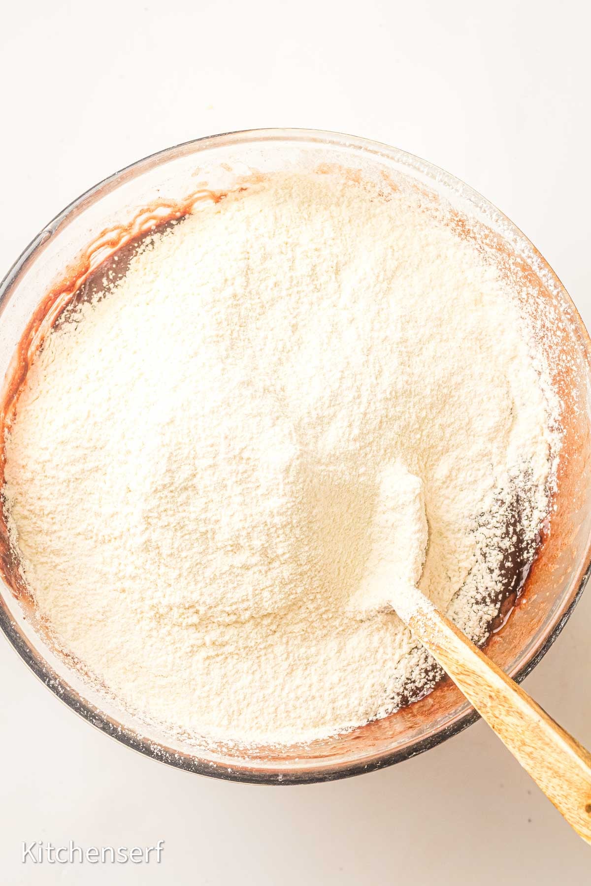 A mixing bowl with flour on top of rich brownies with cocoa powder batter and a wooden spoon partially buried in the flour.