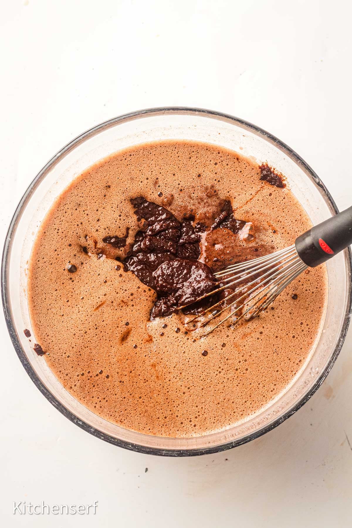 A glass bowl of chocolate batter for brownies with cocoa powder being whisked with a metal whisk on a white surface.