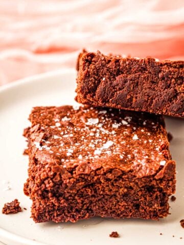 Two thick, fudgy brownies with cocoa powder are topped with flaky sea salt on a white plate, set against a soft pink background.