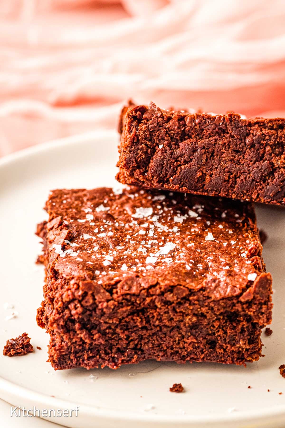 Two thick, fudgy brownies with cocoa powder are topped with flaky sea salt on a white plate, set against a soft pink background.