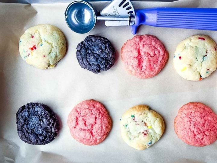 A baking tray with colorful cookies—chocolate, pink, and funfetti cake mix cookies—next to a blue-handled cookie scoop.