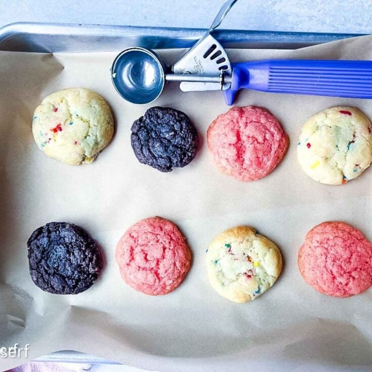 A baking tray with colorful cookies—chocolate, pink, and funfetti cake mix cookies—next to a blue-handled cookie scoop.