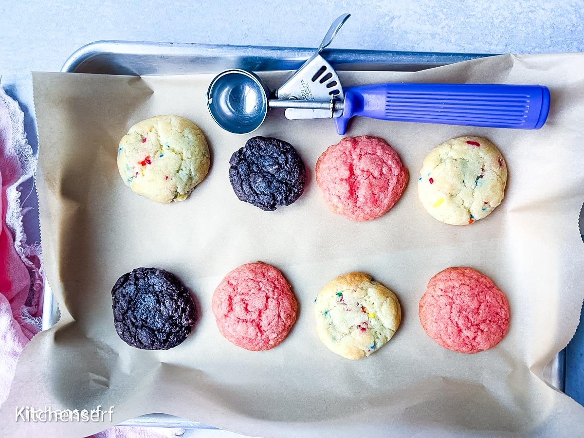 A baking sheet with assorted cake mix cookies and a blue-handled cookie scoop on parchment paper.
