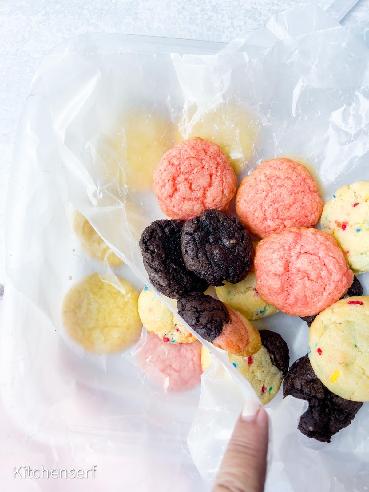 A hand points to a container with yellow, pink, chocolate, and sprinkle cake mix cookies arranged on wax paper.
