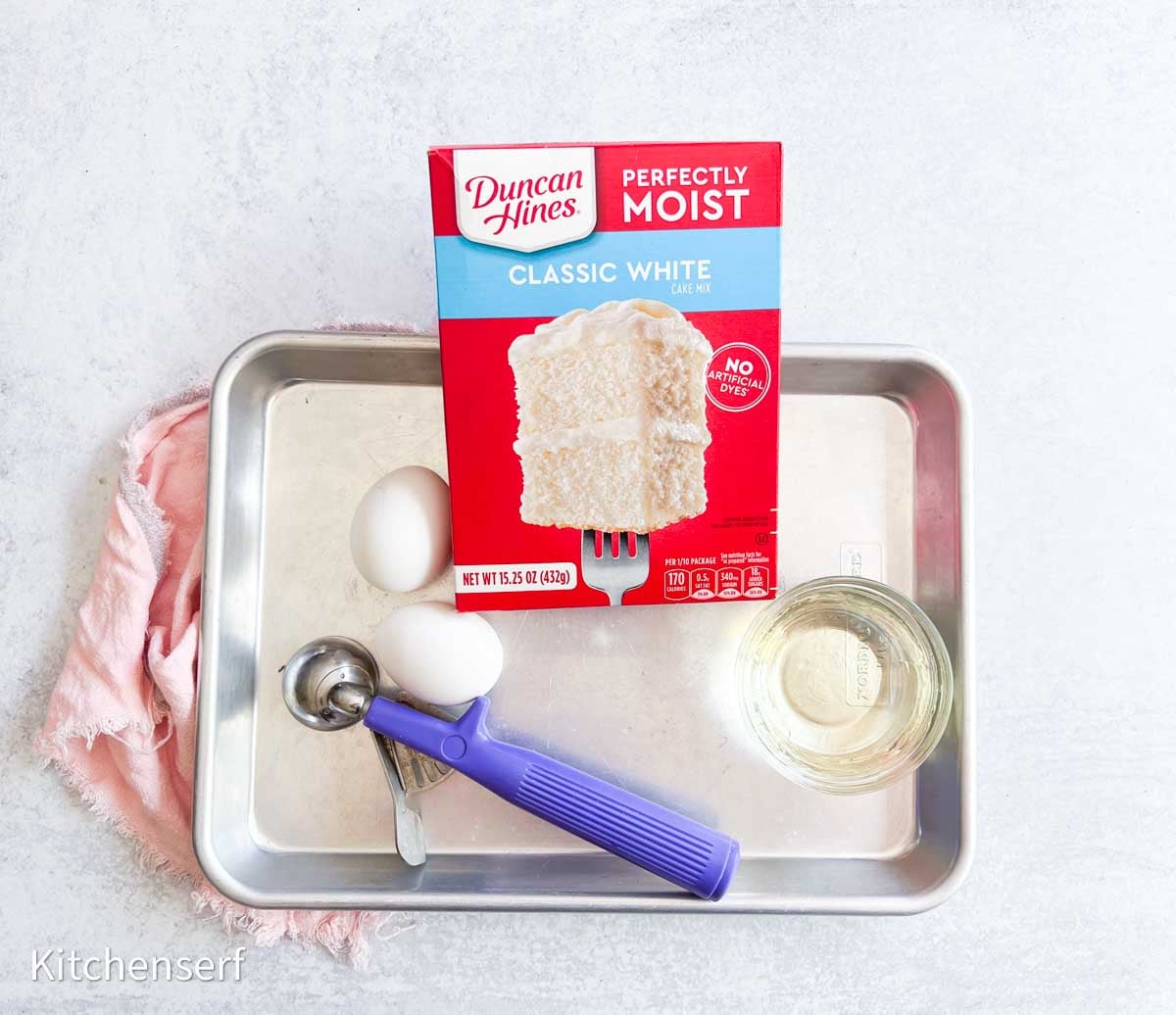 A box of white cake mix, two eggs, oil, and baking tools on a metal tray with a pink cloth underneath—perfect for making cake mix cookies.
