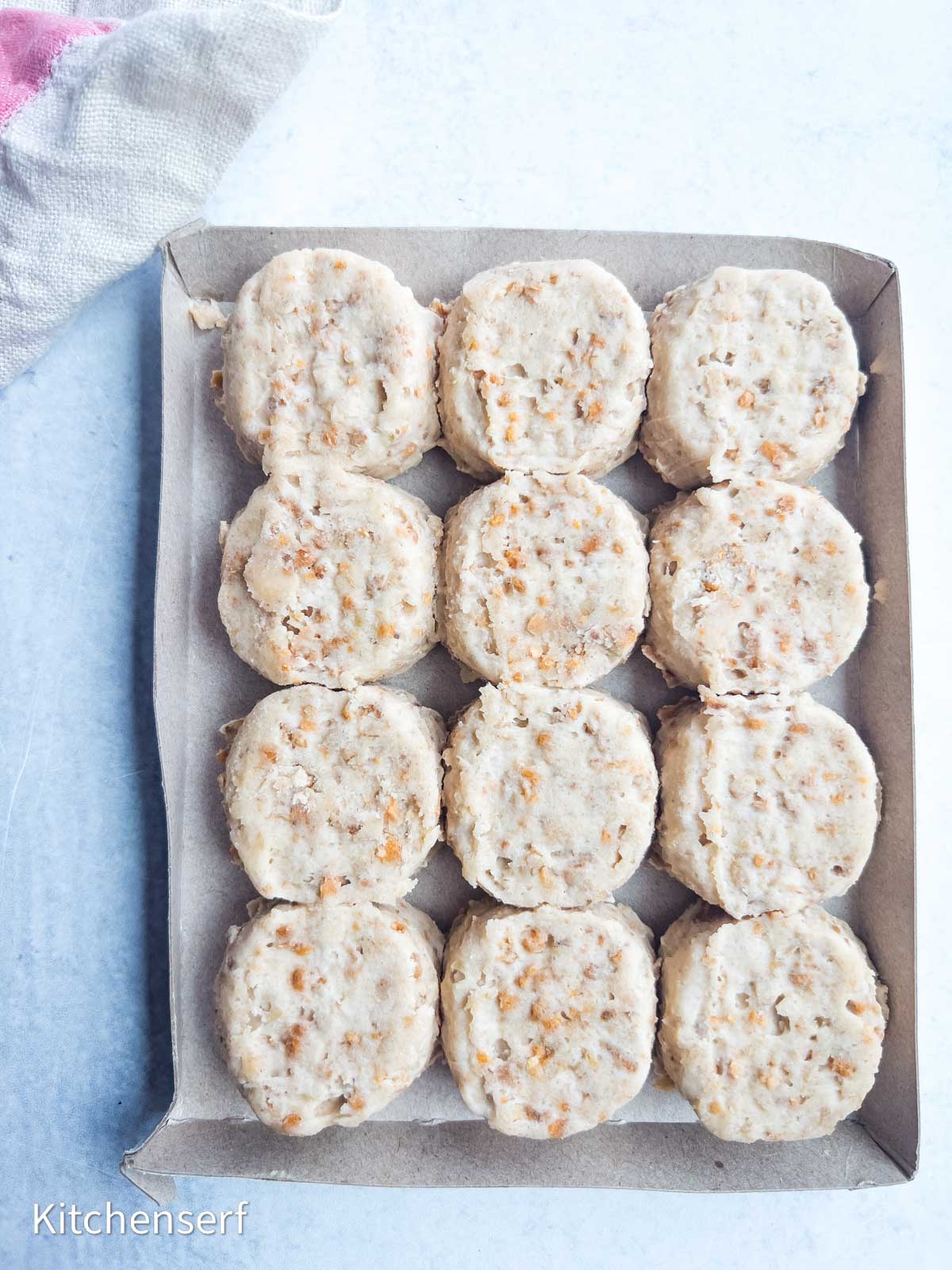 A cardboard tray filled with twelve round, unbaked cheese cookies on a light surface.